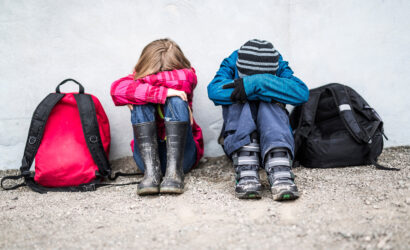Two children sitting on the ground besides their backpacks with their heads down.